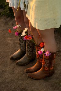 Close-up of floral-filled cowboy boots and dresses in a sunlit field.